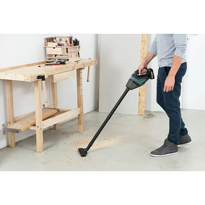 Bosch Power for All 18V Aspirador de mano de batería UniversalVac 18 - 18 V, Sin batería, Volumen de recipiente: 0,5 l - Una persona usa una aspiradora inalámbrica Bosch negra y verde para limpiar virutas de madera del suelo de un taller.