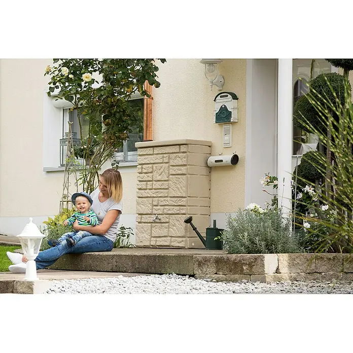 Arves Regenspeicher Maurano - 300 l, Sandsteinfarben, 1 Stk. - Regenwassertank in Steinoptik mit Wasserhahn und Gießkanne, neben einer Frau mit Kind. Praktisch und dekorativ für den Garten.