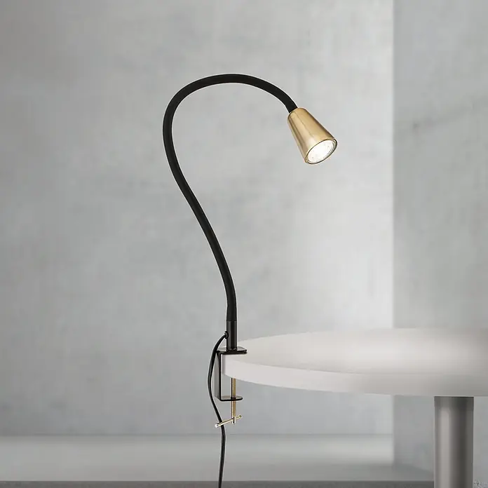 Orion Leuchten Klemmleuchte - 40 W, L x B x H: 7,5 x 24 x 58 cm - Schwarze, flexible Klemmleuchte mit messingfarbenem Kopf, leuchtend an einem weißen Tisch befestigt.