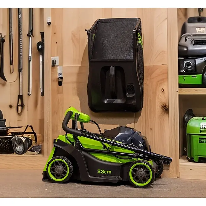 Greenworks Akku-Rasenmäher Gen2 GW 24V BL 4 AH 33 cm - 24 V, 1 Akku, 4 Ah, Schnittbreite: 33 mm - Grüner und schwarzer Elektromäher mit 33cm Schnittbreite, in einem Lagerbereich. Darüber hängt ein schwarzer Grasfangsack.