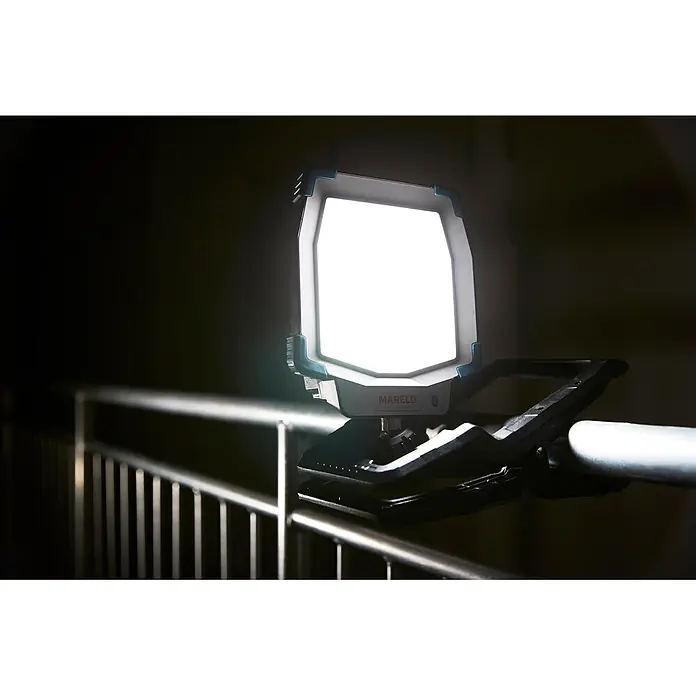 Mareld LED-Strahler Shiny 5000 - 50 W, 5.000 lm, 6.500 K, L x B x H: 28,6 x 18 x 10,2 cm, IP65 - MARELD Arbeitsleuchte, dunkelgrau mit blauen Akzenten, an einem Metallgeländer montiert, beleuchtet einen dunklen Raum.