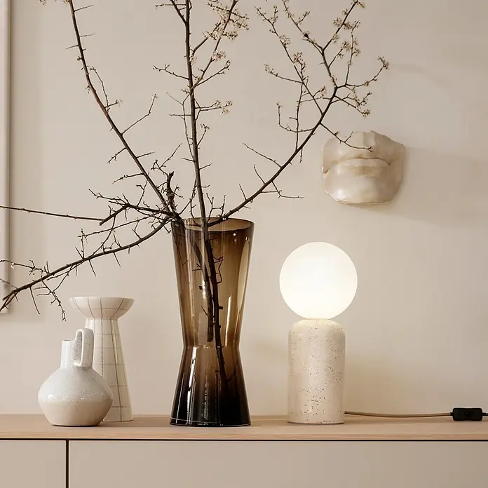Just Light Tischleuchte Traventa - 10 W, L x B x H: 15 x 15 x 35 cm, Travertin, Opalglas, GU10 - Moderne Wohndekoration: Eine braune Glasvase mit Ästen, weiße Keramikgefäße und eine Kugellampe auf strukturiertem Sockel.