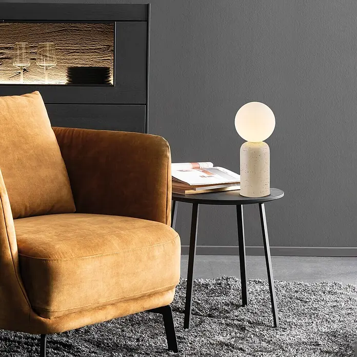 Just Light Tischleuchte Traventa - 10 W, L x B x H: 15 x 15 x 35 cm, Travertin, Opalglas, GU10 - Moderne Tischleuchte mit gesprenkeltem, hellem Sockel und kugelförmiger Leuchtkugel, auf einem Beistelltisch neben einem gelben Sessel.