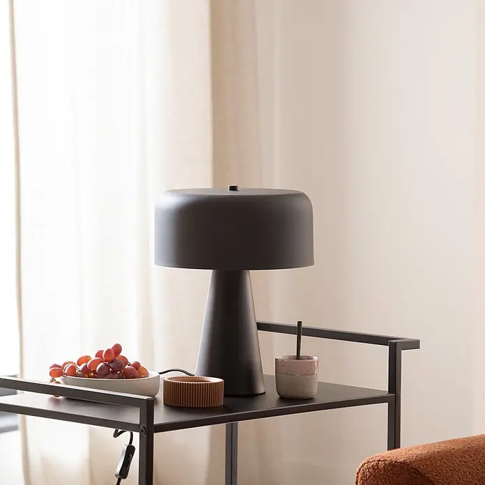 Just Light Tischleuchte Pento - 25 W, L x B x H: 26,5 x 265 x 34,5 cm, Schwarz, E27 - Eine matte schwarze Tischlampe mit breitem, flachem Schirm und konischem Fuß steht auf einem schwarzen Metallregal in einem Wohnzimmer.