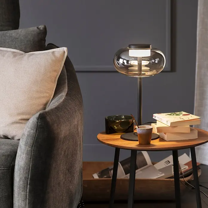 Just Light Akku-Tischleuchte Sia - L x B x H: 22 x 20 x 33 cm, Warmweiß, 9 W, IP20 - Elegante Tischleuchte mit leuchtendem Glasschirm und dunklem Fuß, beleuchtet einen Holznebentisch neben einem gemütlichen Sofa.