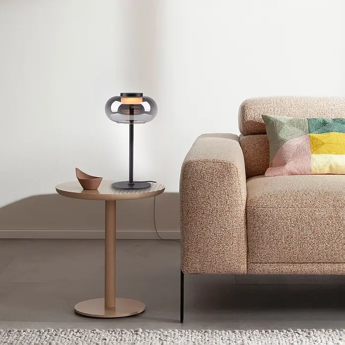 Just Light Akku-Tischleuchte Sia - L x B x H: 22 x 20 x 33 cm, Warmweiß, 9 W, IP20 - Moderne Tischleuchte mit Rauchglasschirm auf einem runden Beistelltisch, neben einem Sofa mit buntem Kissen.