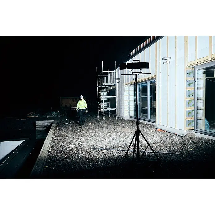 Mareld LED-Strahler Vapor 20000 App & Sensor - 200 W, 20.000 lm, 6.500 K, L x B x H: 43 x 6,7 x 32 cm, IP65 - Starke Arbeitsleuchte auf Stativ beleuchtet nächtliche Baustelle mit Arbeiter in gelber Warnschutzjacke.