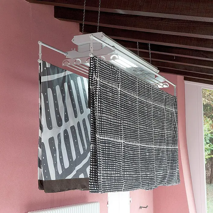 Foxydry Deckenwäschetrockner Air 120 - L x B: 57 x 122 cm, Stahlgrau - Weiße Deckenwäscheleine mit integriertem Licht, an der gemusterte Stoffe in einem Raum hängen.