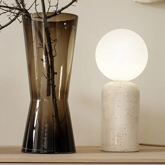 Just Light Tischleuchte Traventa - 10 W, L x B x H: 15 x 15 x 35 cm, Travertin, Opalglas, GU10 - Eine kugelförmige weiße Tischlampe mit cremefarbenem Sockel neben einer hohen, dunkelbraunen Glasvase mit Ästen auf einer Holzoberfläche.