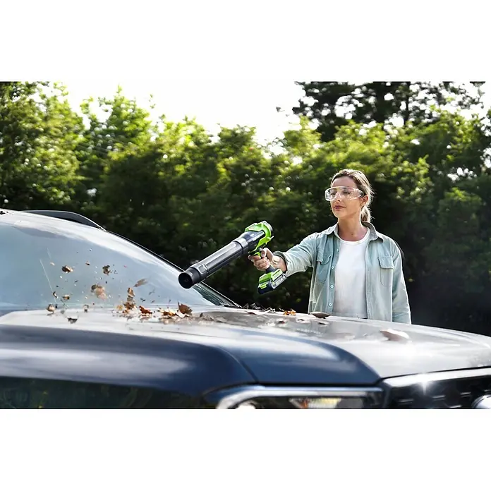 Greenworks Akku-Laubbläser GW 24V BL - 24 V, 1 Akku, 2 Ah, Blasgeschwindigkeit: 209 km/h, Blasleistung: 7,6 m³/h - Frau nutzt grünen Laubbläser, um Blätter von einem dunklen Auto zu entfernen, trägt Schutzbrille.