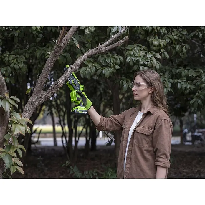 Greenworks Einhand-Akku-Kettensäge Mini GW 24V BL 10 cm - 24 V, 2 Ah, Li-Ionen, 1 Akku, Schwertlänge: 10 cm - Frau schneidet Ast mit grüner und schwarzer Mini-Kettensäge, trägt Schutzbrille und Handschuhe.
