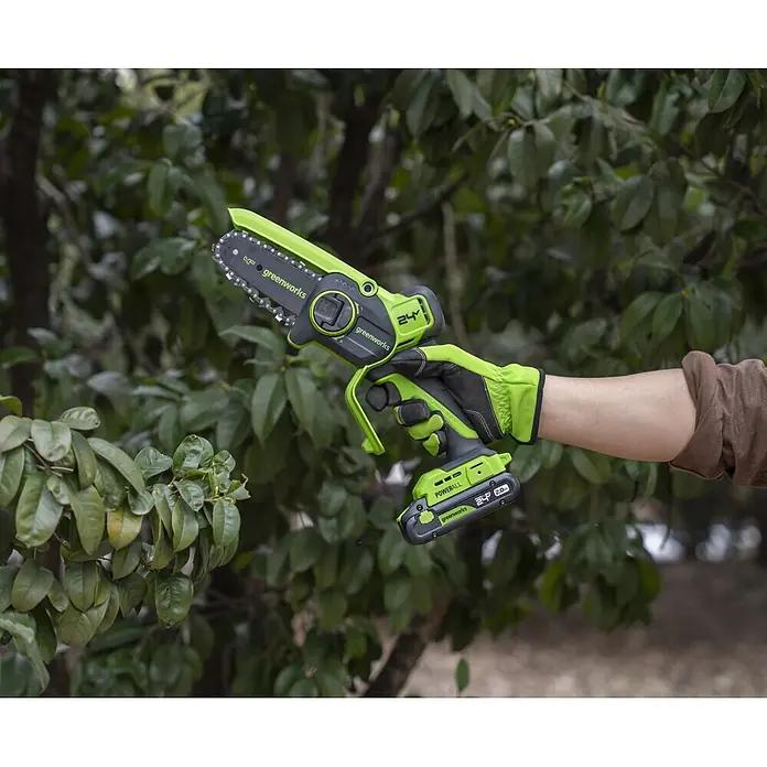 Greenworks Einhand-Akku-Kettensäge Mini GW 24V BL 10 cm - 24 V, 2 Ah, Li-Ionen, 1 Akku, Schwertlänge: 10 cm - Grüne und schwarze Greenworks Mini-Kettensäge, akkubetrieben, von behandschuhter Hand zum Trimmen gehalten.