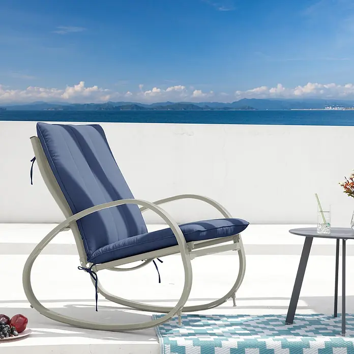 SVITA Garten-Schaukelstuhl EIBY DELUXE - 1 Stk., 50 % Polyester, 50 % Stahl - Outdoor-Schaukelstuhl mit hellem Gestell und blauen Kissen auf einer Terrasse mit Meerblick.