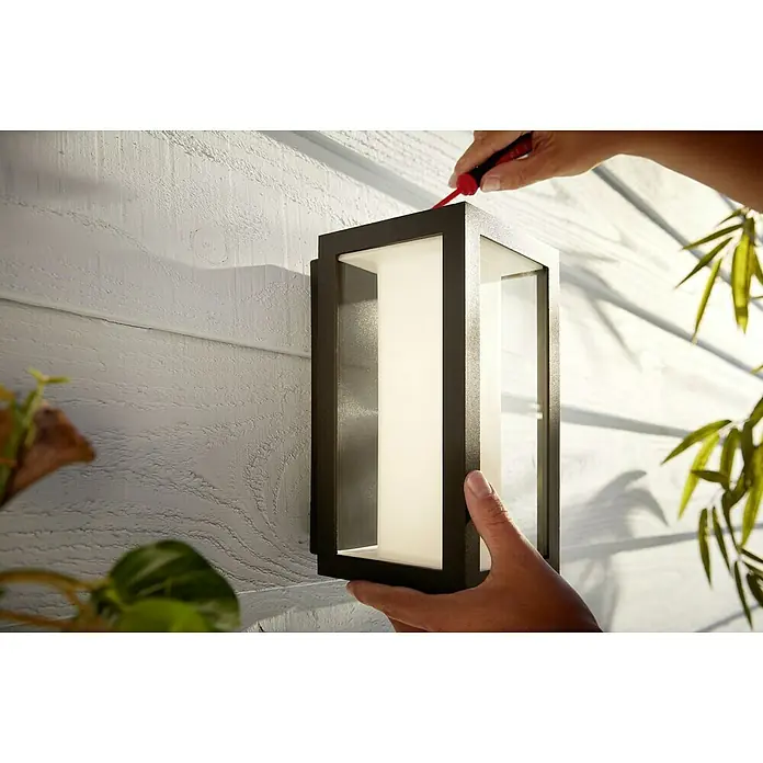 Philips Hue LED-Außenwandleuchte Impress - 8 W, 14,1 x 12 x 24 cm, Schwarz, IP44 - Montage einer modernen Außenleuchte mit Schraubenzieher an einer weißen Holzwand. Die Leuchte hat ein schlankes, rechteckiges Design.