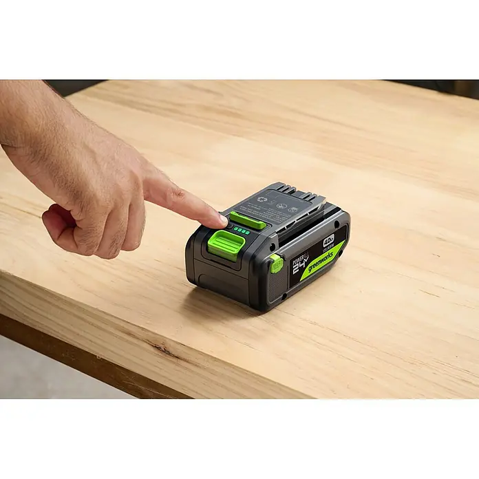Greenworks Akku & Ladegerät 24V 4 AH, VD - Akkuspannung: 24 V, Li-Ionen, 4 Ah - Hand drückt Taste an Greenworks 24V 4.0Ah Akku, drei grüne Ladeanzeigen leuchten, auf Holzoberfläche.