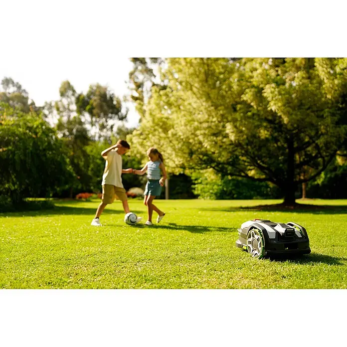 Greenworks Mähroboter AI Mowbot 10 GW KI RTK+VISION - 1.000 m², Schnittbreite: 20 cm, Satellitengestützte Navigation, RTK - Rasenroboter, schwarz-weiß mit grünen Rädern, Kinder spielen im unscharfen Hintergrund.