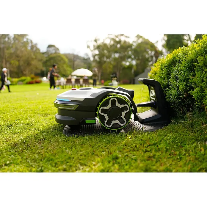 Greenworks Mähroboter AI Mowbot 10 GW KI RTK+VISION - 1.000 m², Schnittbreite: 20 cm, Satellitengestützte Navigation, RTK - Grau-schwarzer Mähroboter mit leuchtend grünen Rädern, der auf einem Rasen neben einem Busch lädt.