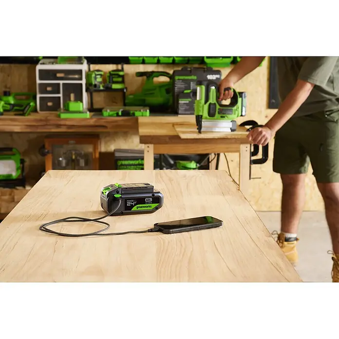 Greenworks Akku & Ladegerät 24V 4 AH, VD - Akkuspannung: 24 V, Li-Ionen, 4 Ah - Greenworks 24V 4.0Ah POWERALL Akku, schwarz-grün, lädt ein Smartphone auf einer Werkbank, mit Person, die ein Nagelgerät nutzt, im Hintergrund.
