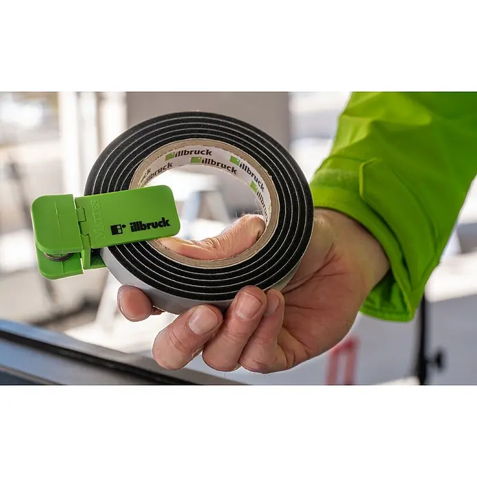 Illbruck Fugendichtband TP670 ML-Tech Pro - Fugenbreite: 6 mm - 18 mm, Fugentiefe: 20 mm, Anthrazit, Länge: 5,3 cm, Passend für: Fenster- und Türenmontage - Eine Person hält eine Rolle schwarzes illbruck Dichtband mit einem grünen MATTES Abroller, bereit zur Anwendung.