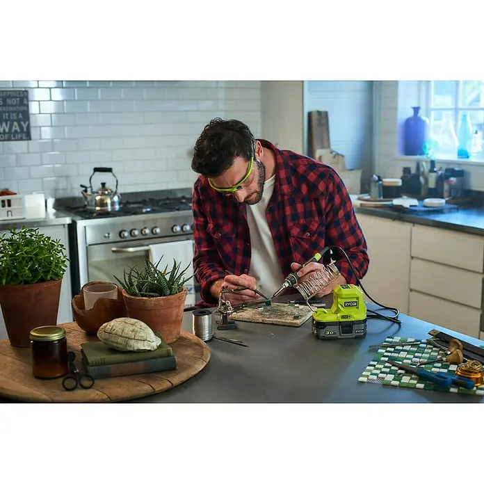 Ryobi ONE+ Soldador de puntos de batería R18SOI-0 - Apto para: Soldaduras finas - Un hombre usando un soldador Ryobi en una pieza de mosaico, con herramientas y plantas en la mesa.