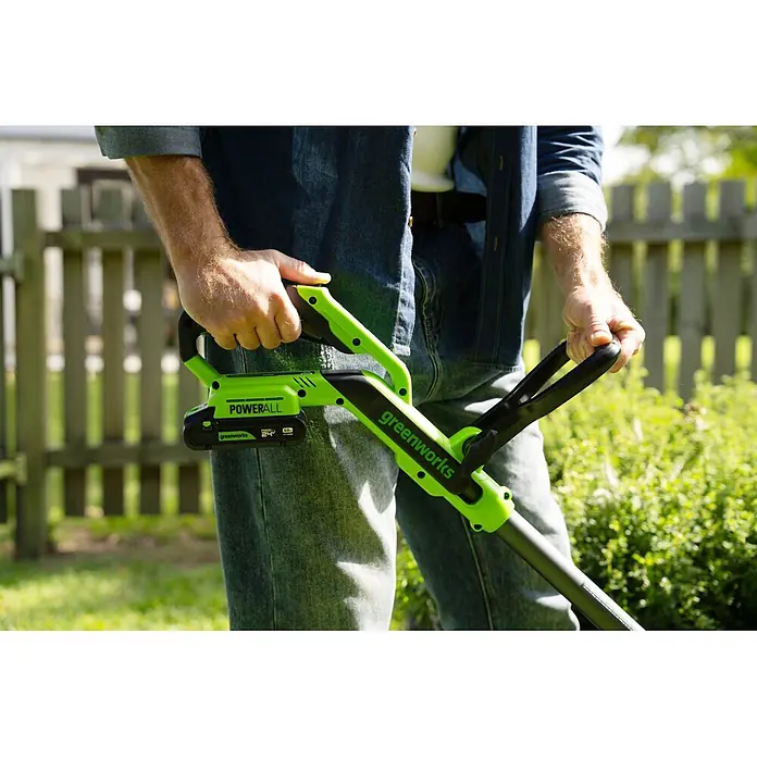 Greenworks Akku-Rasentrimmer 24V BL 30 cm - 24 V, 2 Ah, Li-Ionen, 1 Akku, Schnittbreite Messer: 30 cm, Schnittbreite Faden: 30 cm - Eine Person hält einen grünen und schwarzen Greenworks POWERALL Akku-Rasentrimmer mit sichtbarem Akku, bereit für den Außeneinsatz.