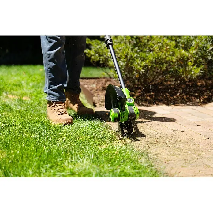 Greenworks Akku-Rasentrimmer 24V BL 30 cm - 24 V, 2 Ah, Li-Ionen, 1 Akku, Schnittbreite Messer: 30 cm, Schnittbreite Faden: 30 cm - Grüner Rasentrimmer schneidet Gras am gepflasterten Wegrand; Grasschnitt fliegt umher.