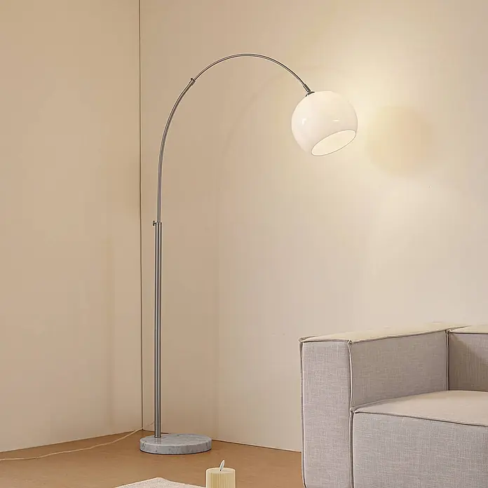 Lindby Stehleuchte Alverton - Höhe: 197 cm, alu / grau / zink, weiß / opal, E27 - Bogenförmige Stehleuchte mit silbernem Gestell, weißem Kugelschirm und Marmorfuß, neben einem Sofa.