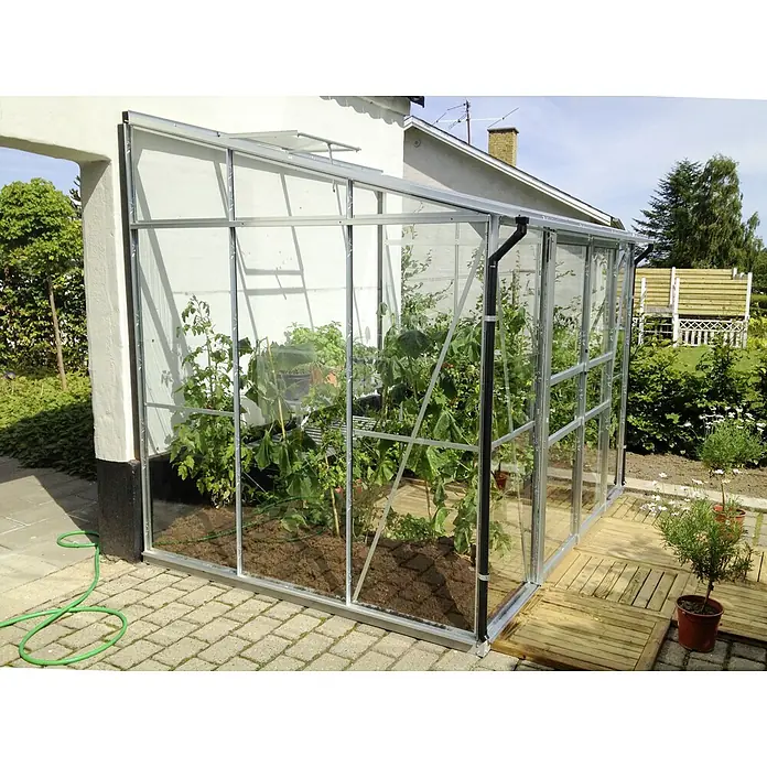 Vitavia Gewächshaus Osiris 5200 - Außenmaß inkl. Dachüberstand (B x T): 262,1 x 201 cm, 3 mm, Aluminium - Dieses Bild zeigt ein Gewächshaus mit Pflanzen im Inneren, das an ein Gebäude angebaut ist. Das Gewächshaus ist aus Metall und Glas gefertigt.