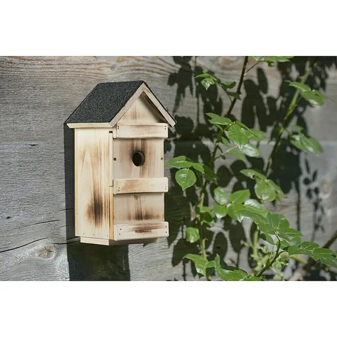 Windhager Nistkasten - Geeignet für: Meisen, L x B x H: 15 x 16 x 31 cm, Massivholz, Geflammt - Das Bild zeigt ein Vogelhaus aus Holz mit einem dunklen Dach, das an einer Holzwand befestigt ist und von Blättern umgeben ist.