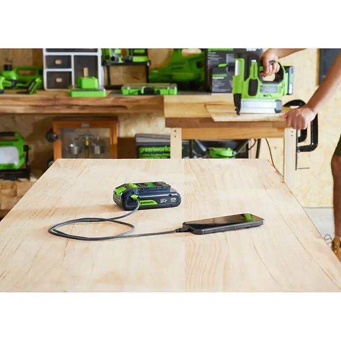 Greenworks Akku & Ladegerät 24V 2 AH, VD - Akkuspannung: 24 V, Li-Ionen, 2 Ah - Ein schwarzer und grüner Greenworks PowerAll 24V Akku (2.0Ah USB-C 65W) lädt ein Smartphone auf einer Holzwerkbank.
