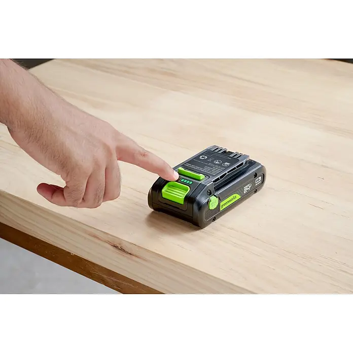Greenworks Akku & Ladegerät 24V 2 AH, VD - Akkuspannung: 24 V, Li-Ionen, 2 Ah - Eine Hand drückt den Ladezustands-Knopf einer schwarz-grünen Greenworks 24V Batterie, die ihren Ladezustand auf Holz anzeigt.