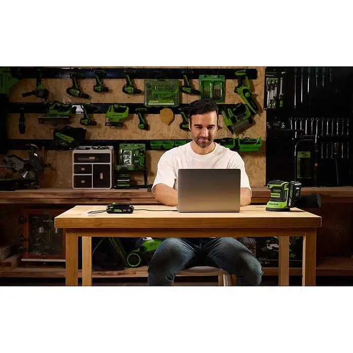 Greenworks Akku & Ladegerät 24V 2 AH, VD - Akkuspannung: 24 V, Li-Ionen, 2 Ah - Mann benutzt Laptop, verbunden mit einer Greenworks Powerbank, auf einem Werkstatttisch mit Schleifer und weiteren Werkzeugen.