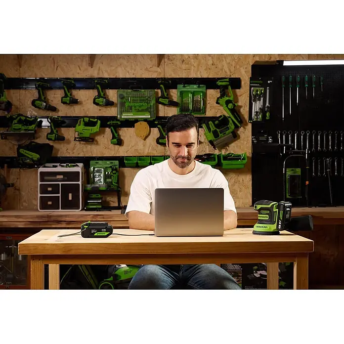 Greenworks Akku 24V 4 AH HP mit USB-C - 24 V, 4 Ah, Li-Ionen - Mann nutzt grüne und schwarze Greenworks Batterie als Powerbank für seinen Laptop, mit passendem Schleifer auf der Werkbank voller Werkzeuge.