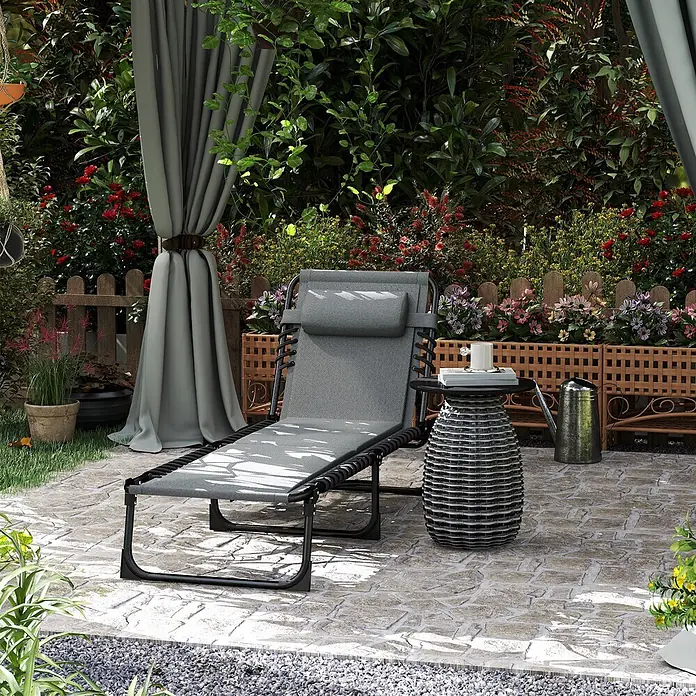 Furnicato Gartenliege - Dunkelgrau, Polyester - Graue Outdoor-Liege mit passender Kopfstütze und Beistelltisch aus Korbgeflecht auf einer Steinterrasse in einem üppigen Garten.