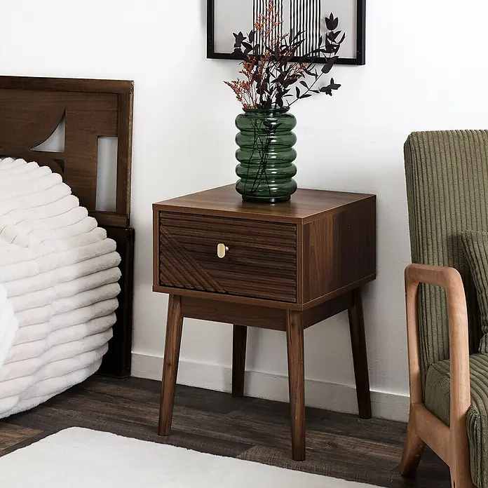 Home Deco Factory Tiffany Mesita de noche - L x An x Al: 40 x 40 x 55 cm, Marrón, Número de cajones: 1 ud. - Mesita de noche de madera oscura con cajón texturizado y pomo dorado, junto a una cama con un jarrón.