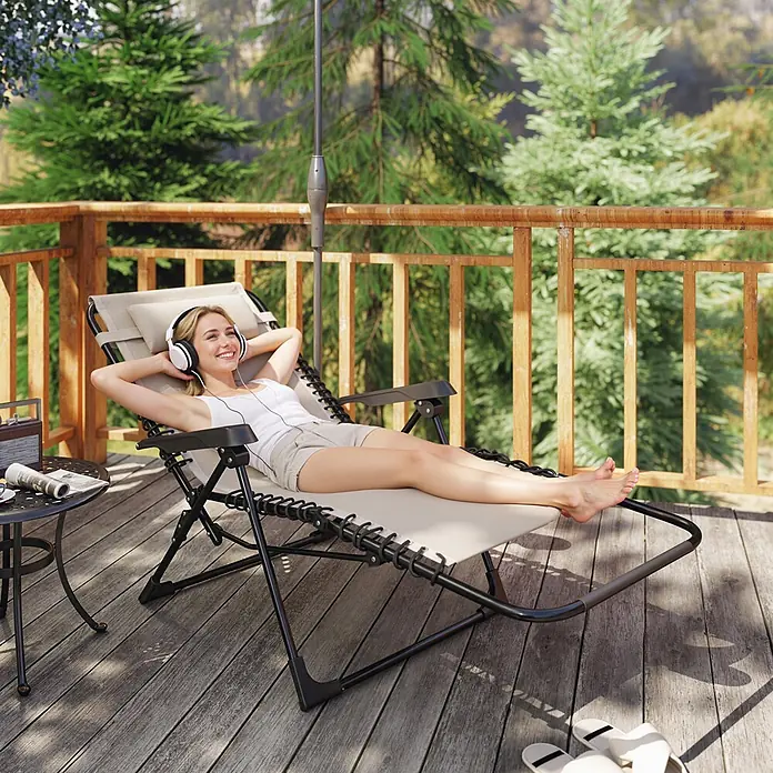 Outsunny Garten-Liegestuhl - Metall, Textelin, Klappbar, Beige, B x T x H: 65 x 160 x 112 cm - Eine Frau entspannt in einem beigefarbenen Zero-Gravity-Liegestuhl mit schwarzem Gestell auf einer Holzterrasse im Freien.