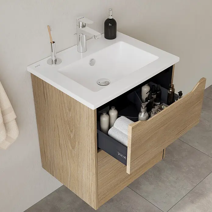 VitrA Integra Square Badmöbel-Set - Gesamtbreite Badmöbel: 60 cm, Eiche Gold - Badwaschtischunterschrank aus hellem Holz mit weißem Waschbecken und offener Schublade, gefüllt mit Pflegeprodukten und Handtüchern.