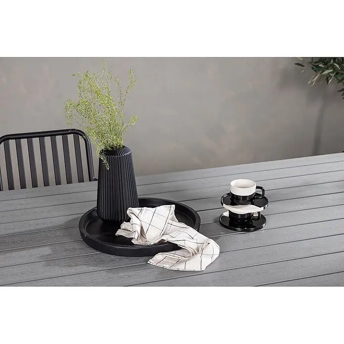 Ebuy24 Gartentisch Garcia - L x B x H: 200 x 100 x 74 cm, Ø x H: 100 x 74 cm, Nonwood, Aluminum, Grau - Eine schwarze gerippte Vase mit Grünpflanzen, ein schwarzes rundes Tablett mit karierter Serviette und gestapelte schwarz-weiße Tassen auf einem grauen Gartentisch.