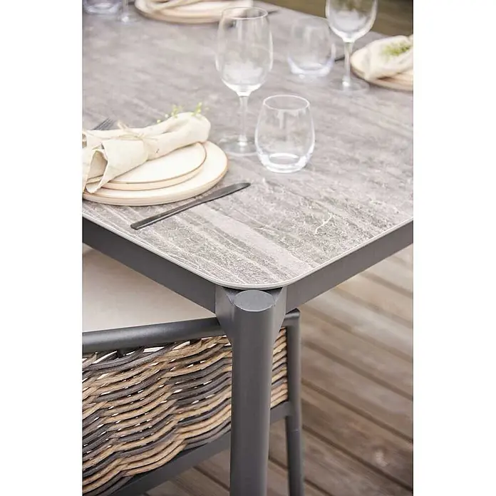 Ebuy24 Gartentisch Bo - L x B x H: 104 x 205 x 75 cm, Ø x H: 205 x 75 cm, Aluminium Komposit, Sonstige, Schwarz - Ecke eines Outdoor-Esstisches mit grauer Marmorplatte und dunkelgrauem Gestell, neben einem Stuhl mit geflochtener Rückenlehne, auf einer Holzterrasse.
