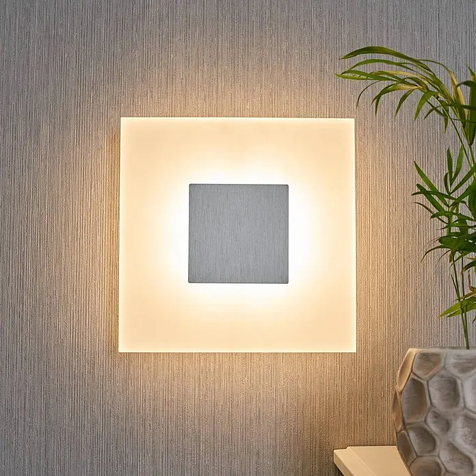 Lucande Wandleuchte Berlind - L x B x H: 6 x 26 x 26 cm, weiß / opal - Quadratische Wandleuchte mit gebürsteter Metallmitte, die warmes Licht auf eine strukturierte Wand wirft, neben einer Pflanze.