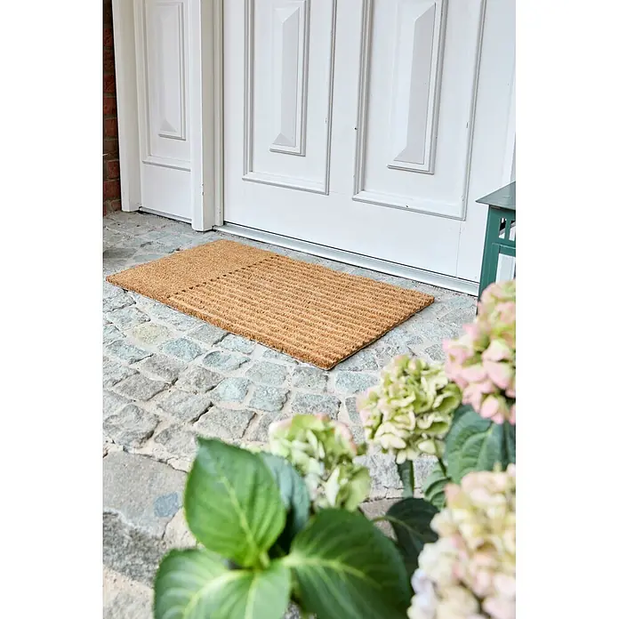 Fußmatte Coco Shape - 75 x 45 cm, Natur, Material Nutzschicht: 100 % Kokos, Streifen - Eine natürliche Kokos-Fußmatte mit gestreiftem Muster vor einer weißen Tür auf einem Kopfsteinpflasterweg.