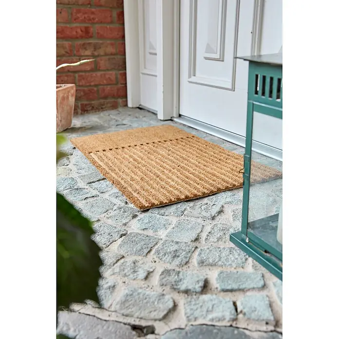 Fußmatte Coco Shape - 75 x 45 cm, Natur, Material Nutzschicht: 100 % Kokos, Streifen - Braune Kokosfasermatte mit gerippter Textur, auf Steinpflaster vor einem Hauseingang platziert.