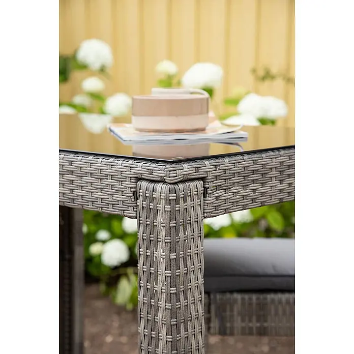 Ebuy24 Gartentisch Alo - L x B x H: 100 x 60 x 110 cm, Ø x H: 60 x 110 cm, Glas, Stahl, Korbmöbel, Sonstige, Grau - Nahaufnahme eines grauen, geflochtenen Rattan-Gartentisches mit Glasplatte, darauf eine Tasse und Zeitschriften. Teil eines Outdoor-Sets.