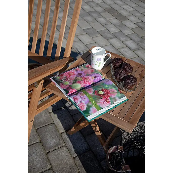 Ebuy24 Gartentisch Bine - L x B x H: 50 x 50 x 50 cm, Ø x H: 50 x 50 cm, Teakholz massiv, Sonstige, Braun - Hölzerner Outdoor-Beistelltisch mit einem aufgeschlagenen Buch, einer Tasse und Muffins, neben einem Holzstuhl.