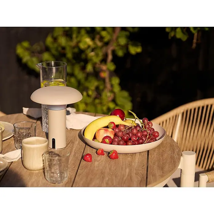 Ebuy24 Gartentisch Bella - L x B x H: 90 x 90 x 74 cm, Ø x H: 90 x 74 cm, Nonwood (PE), Aluminium, Sandfarben - Beigefarbene, pilzförmige, tragbare Tischlampe auf einem Holztisch im Freien mit Obst und Getränken.