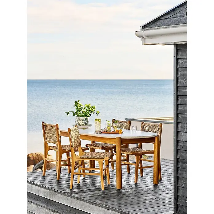 Ebuy24 Gartentisch Tirsa - L x B x H: 95 x 210 x 76 cm, Ø x H: 210 x 76 cm, Akazie, Terrazzo, Sonstige, Natur - Outdoor-Essgruppe: ovaler Tisch mit heller Platte, vier Holzstühle mit geflochtenen Sitzen, auf einer Terrasse am Meer.