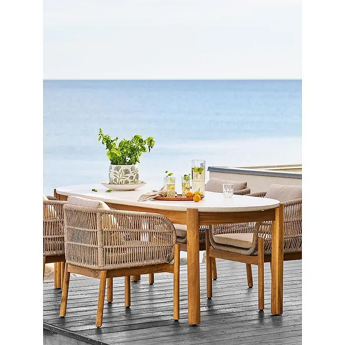 Ebuy24 Gartentisch Tirsa - L x B x H: 95 x 210 x 76 cm, Ø x H: 210 x 76 cm, Akazie, Terrazzo, Sonstige, Natur - Eine Outdoor-Essgruppe mit weißem Ovaltisch und geflochtenen Stühlen mit hellen Kissen auf einer Holzterrasse am Meer.
