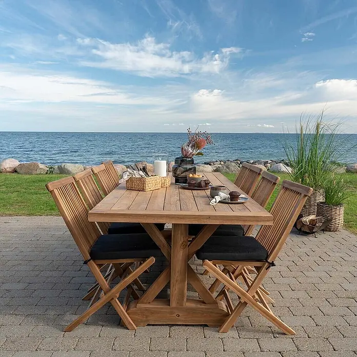 Ebuy24 Gartentisch Volos - L x B x H: 240 x 100 x 75 cm, Ø x H: 100 x 75 cm, Teakholz, Teak - Teakholz-Gartenessgruppe mit 8 Stühlen und schwarzen Kissen auf einer Terrasse am Meer.