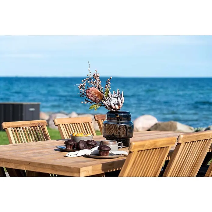 Ebuy24 Gartentisch Volos - L x B x H: 240 x 100 x 75 cm, Ø x H: 100 x 75 cm, Teakholz, Teak - Hölzernes Outdoor-Essgruppe mit Lattenstühlen und Tisch, mit Snacks und Vase am Meer.