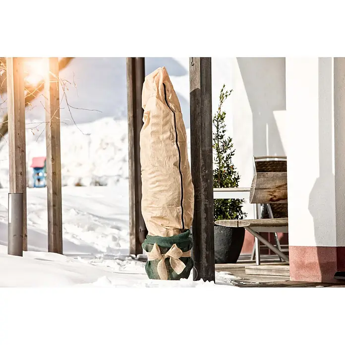 Windhager Haubenvlies - B x H: 0,6 x 1,8 m, Beige, 50 g/m² - Beiger Pflanzenschutzbeutel mit grünem Jute-Unterteil und Schleife, im Schnee stehend.